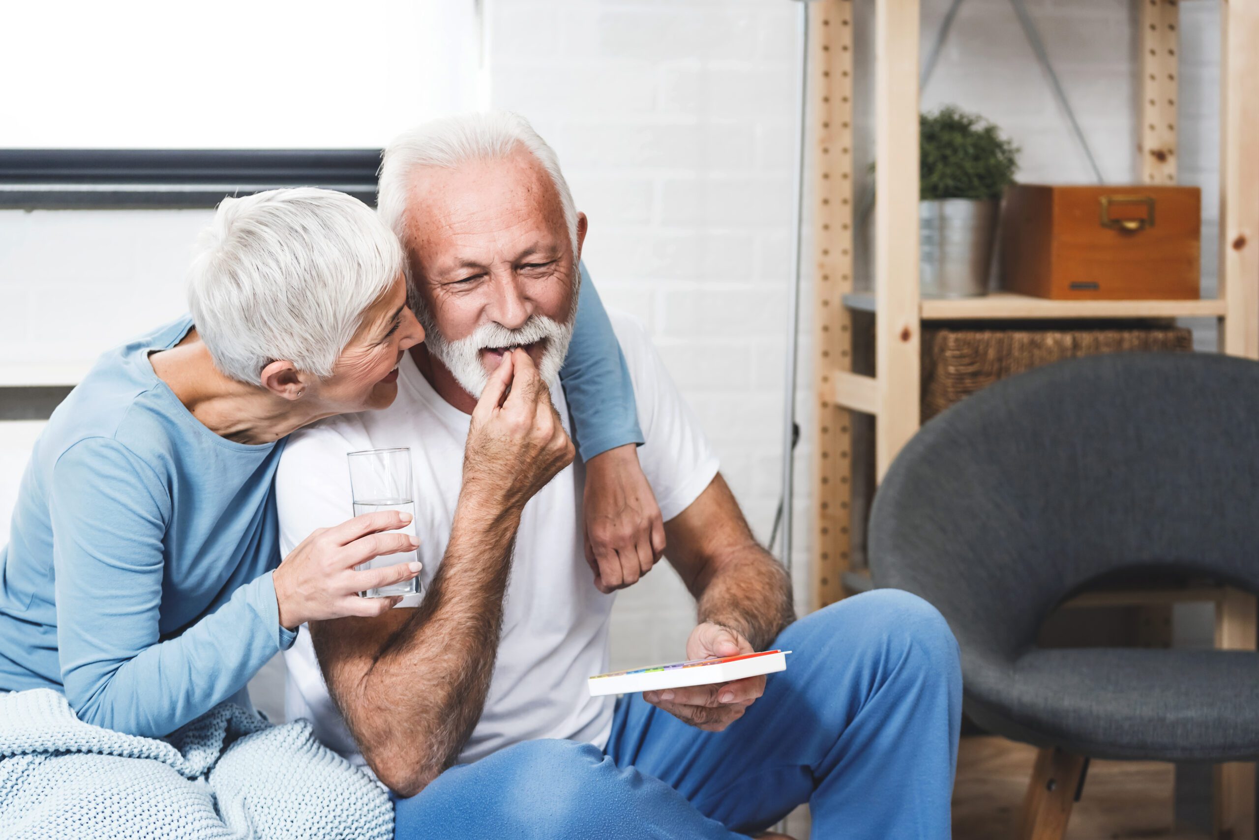 Senior man laughing while take medication for potency while his wife smile too. Elderly couple in bedroom.