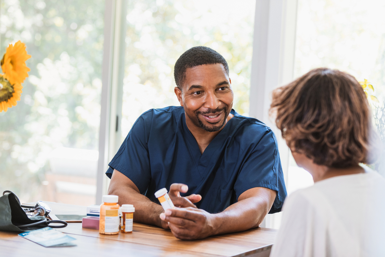 Sitting at the senior woman's dining room table the mid adult home health nurse explains the medication to his senior patient.