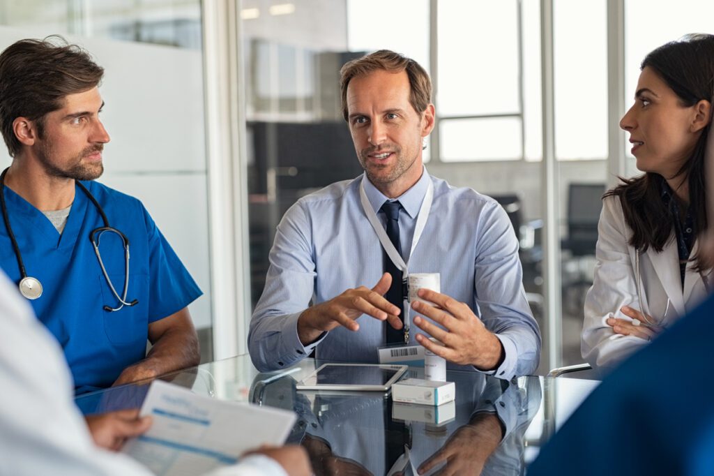 Mature man with professional doctors in hospital showing medicine bottles. Confident representative pharmaceutical showing new medicines to a team of doctor and nurse. Specialist showing bottle of pills and explaning drug dosage to a group of specialist in meeting room.