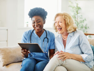 Doctor or nurse caregiver showing a tablet screen to senior woman and laughing at home or nursing home