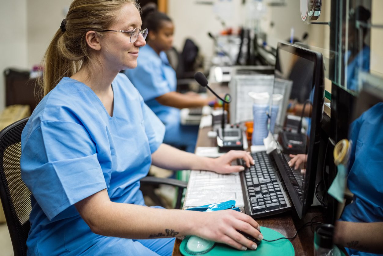 Two nurses using computers, sitting in reception in medical clinic.