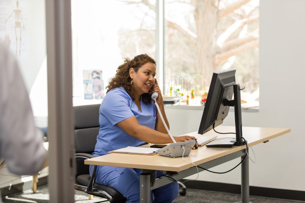 While talking to a patient on the telephone, the mid adult female nurse refers to information on the screen of the desktop PC.