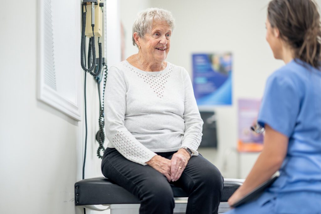 A senior woman sits up on an exam table during a routine check-up. She is dressed casually and listening attentively to her female doctor who is seated in front of her in blue scrubs.