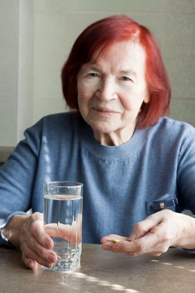 elderly woman holding pills and glass of fresh water, takeing medicines or vitamins. Blurred portrait of sad old woman with red hair sitting at table at home. Focus on pills. Medicine, health care