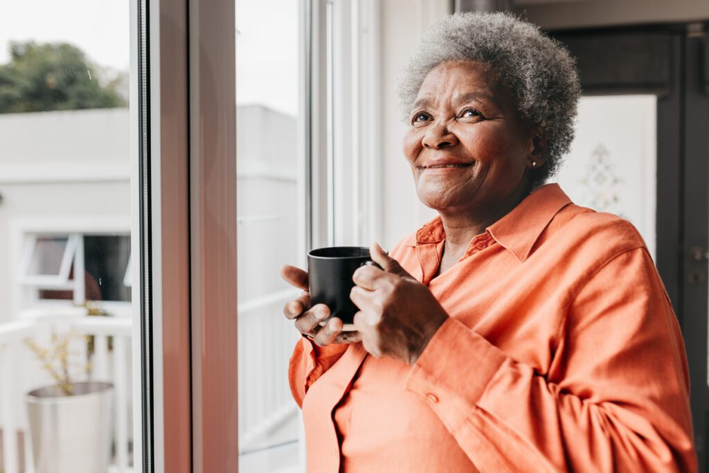 Senior woman holding mug, smiling and looking up, standing beside window at home