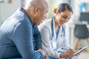 A female doctor of mixed race, sits casually beside a senior male patient in a waiting room, with a tablet in hand, as they review and fill out some intake forms together.