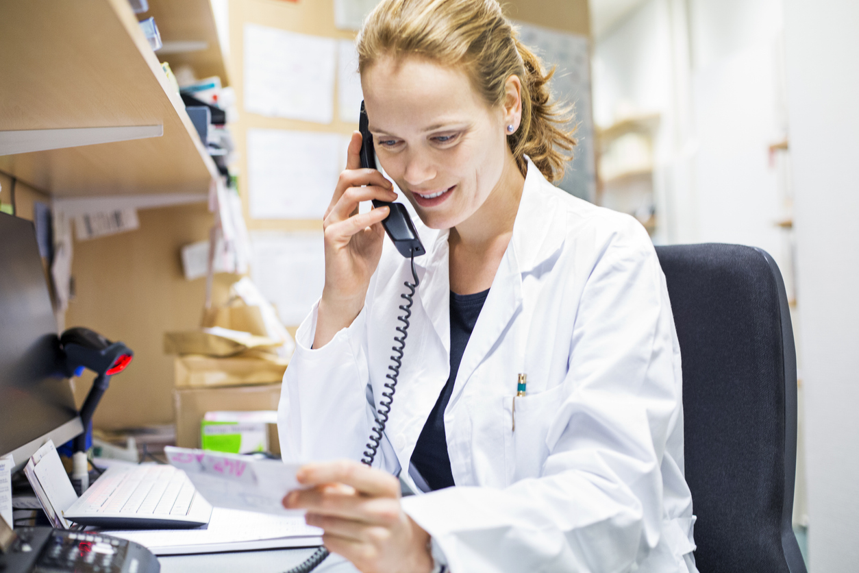 Smiling female pharmacist using landline phone in store. Chemist is reading prescription while sitting at desk. She is working at pharmacy.