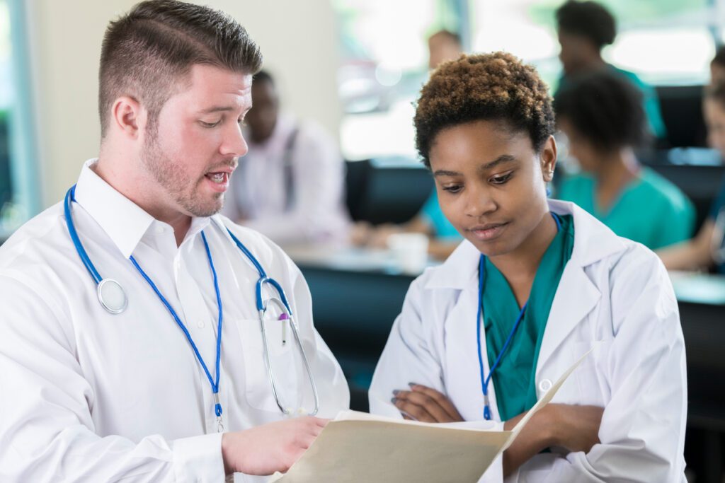 A male guest speaker discusses his upcoming lecture in a medical training class with the professor. She stands in the classroom and listens with arms folded.